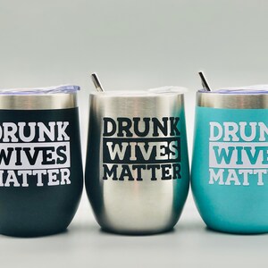 May include: Three stainless steel wine tumblers with lids and straws. The tumblers are black, silver, and light blue. Each tumbler has the text "Drunk Wives Matter" printed on it.