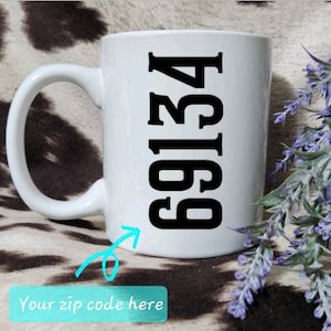 May include: White ceramic mug with a large black number 69134 printed vertically. The mug has a curved handle and is set against a background with purple flowers and a cowhide pattern.