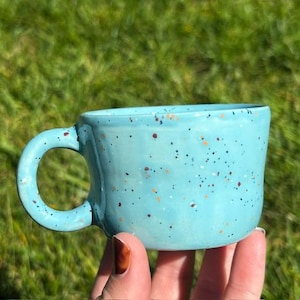 May include: A light blue ceramic mug with a glossy finish and a speckled pattern featuring small brown, orange, and black dots. The mug has a rounded handle and a slightly irregular, organic shape, showcasing a smooth, glazed surface texture.
