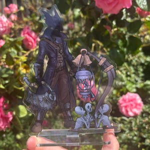 May include: A clear acrylic figure featuring a character in a dark blue coat, holding a lantern and a weapon. The figure has a wolf-like head and is accompanied by two small, pale figures. The background is a garden with pink roses.