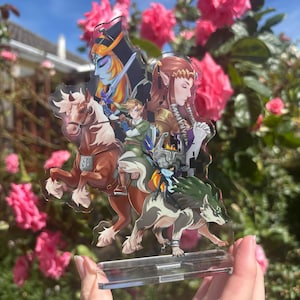 May include: A clear acrylic stand featuring a colorful illustration of characters from a video game. The artwork includes a knight on a horse, a wolf, and other figures. The background is a blur of pink flowers and a blue sky.