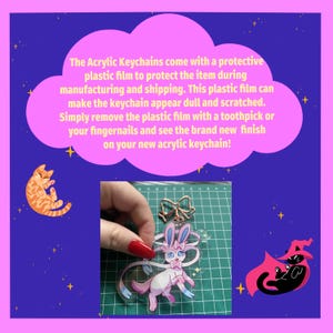 May include: A clear acrylic keychain featuring a pink cartoon bunny with a blue bow. The keychain is being held by a hand with red nail polish. The keychain is on a green grid background.
