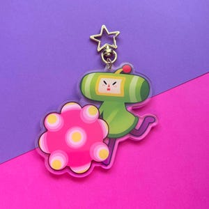 May include: A clear acrylic keychain featuring a green character with a red sphere on its head and a pink object with yellow circles. The keychain has a gold star-shaped clasp. The background is a split of purple and pink.