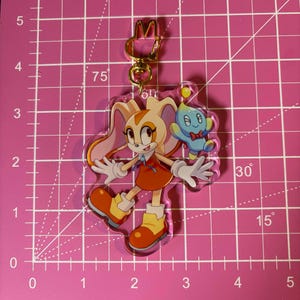 May include: A clear acrylic keychain featuring Cream the Rabbit and a Chao from the Sonic the Hedgehog series. Cream is in a red dress and orange boots, with a blue Chao character. The keychain has a gold-colored clasp.