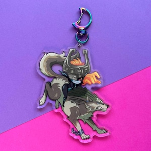 May include: A clear acrylic keychain featuring a cartoon illustration of a wolf-like creature with a female character riding on its back. The female character is wearing a dark green outfit and a large, dark gray helmet with a red eye. The wolf is gray and white with a green mane. The keychain has a silver metal hook.