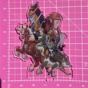 May include: A clear acrylic standee featuring characters from The Legend of Zelda series. The design includes Link on horseback, a wolf, and other characters in a colorful, detailed illustration. The standee is on a pink grid background.
