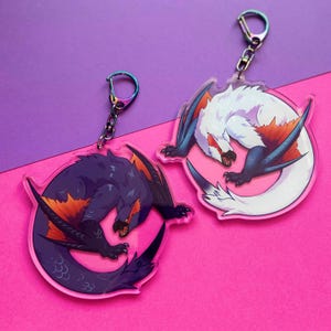May include: Two black and white acrylic keychains featuring a dragon-like creature with orange accents. The creature is curled in a circle with its tail in its mouth. The keychains are on a pink background.