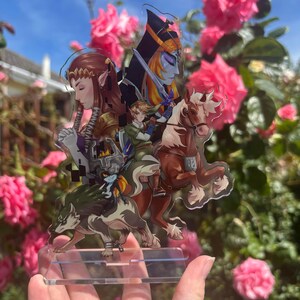 May include: A clear acrylic stand featuring a colorful illustration of characters from a fantasy video game. The artwork includes a knight on horseback, a wolf, and other figures, set against a backdrop of pink roses and a blue sky.