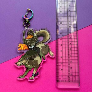 May include: A clear acrylic keychain featuring a cartoon illustration of a wolf and a character from the Legend of Zelda video game series. The character is riding the wolf and holding a sword. The keychain is approximately 6 cm tall.