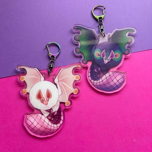 May include: Two acrylic bat keychains. One bat is white with pink wings and a pink tail. The other bat is purple with green wings and a pink tail. Both keychains have a silver keyring.