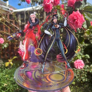 May include: Acrylic stand featuring two anime-style figures, one in a red coat and the other in a black bodysuit. Both figures are holding guns. The stand has a gold and purple circular base with text around the edge.