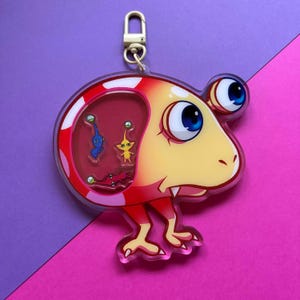 May include: A red and yellow cartoon character keychain with a clear plastic window showing three smaller cartoon characters inside. The larger character has a large, toothy grin and two large eyes.