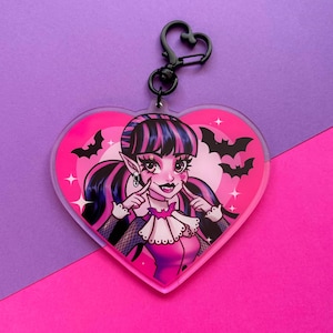 May include: A pink heart-shaped acrylic keychain with a cartoon image of a vampire girl with pink and black hair, wearing a pink and black outfit, and a black choker with a bat charm. The background is pink with white stars and bats.