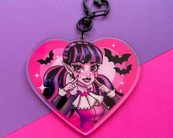May include: A pink heart-shaped acrylic keychain featuring a cartoon image of a vampire girl with pink and black hair, wearing a pink and black outfit, and a black choker with a bat charm. The background is pink with white stars and bats.