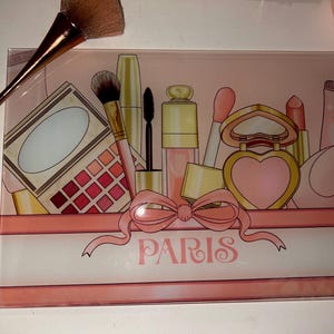 May include: A decorative glass tray with a pink and white design featuring illustrations of makeup products. The word "PARIS" is written in cursive. A large makeup brush is placed on the tray.