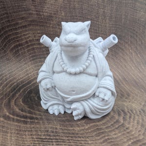 May include: A light gray, speckled figurine of a seated creature with a rounded belly, wearing a beaded necklace. The figure features two cannon-like protrusions extending from its back, pointed ears, and a stoic facial expression with a wide mouth.