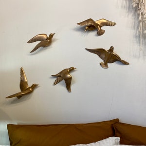 May include: Five gold-colored bird wall sculptures in various flying poses are mounted on a light gray wall. The birds have outstretched wings and are arranged in a circular pattern. A brown pillow and white bedding are visible below.
