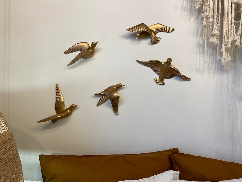 May include: Five gold-colored bird wall sculptures in various flying poses are mounted on a light gray wall. The birds have outstretched wings and are arranged in a circular pattern. A brown pillow and white bedding are visible below.