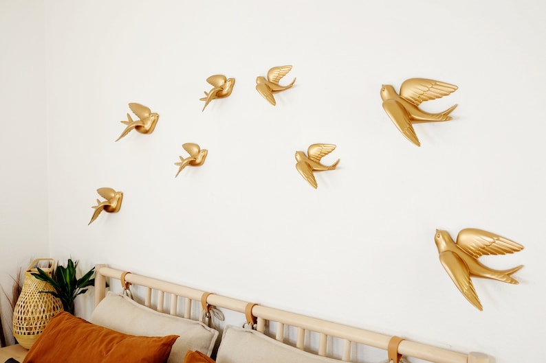 May include: Gold-colored swallow wall decor arranged in a dynamic pattern on a white wall. The swallows are in various stages of flight, adding a sense of movement. The room features a bed with neutral-toned bedding and a woven basket.