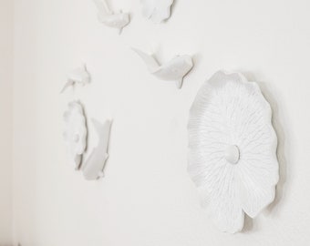 Op de afbeelding: Witte keramische wanddecoratie met een grote lotusbloem en verschillende kleinere visvormen. De decoratie is gemonteerd op een witte muur.