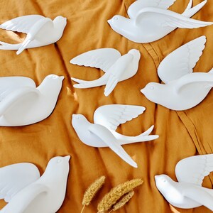 May include: A collection of white swallow bird decorations in various poses, with wings spread in flight. The birds are arranged on a mustard-colored fabric, creating a decorative display. The swallows are made of a smooth material.