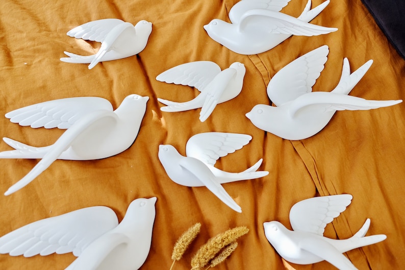 May include: A collection of white swallow bird decorations in various poses, with wings spread in flight. The birds are arranged on a mustard-colored fabric, creating a decorative display. The swallows are made of a smooth material.