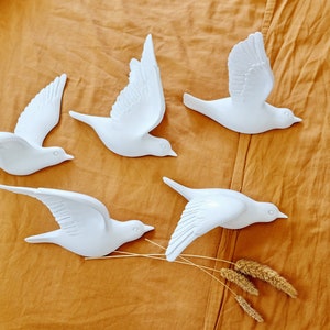 May include: Five white decorative bird sculptures in various flying poses are arranged on a textured, golden-brown surface. The birds have detailed wing and feather designs, and some are accompanied by dried wheat stalks.