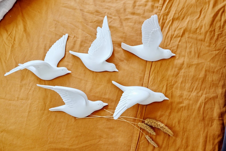 May include: Five white decorative bird sculptures in various flying poses are arranged on a textured, golden-brown surface. The birds have detailed wing and feather designs, and some are accompanied by dried wheat stalks.