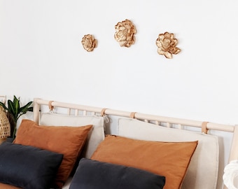Op de afbeelding: Drie gouden, bloemvormige wanddecoraties zijn op een witte muur gemonteerd. Daaronder is een bedframe met een lichte houten afwerking versierd met kussens in zwart, beige en roestkleuren. Het bed is bedekt met een roestkleurige deken.