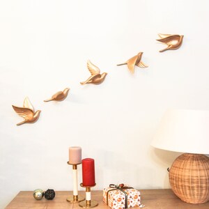 May include: A wall decoration featuring six gold-colored bird sculptures in various stages of flight. The birds are mounted on a white wall above a wooden table. A lamp with a woven base and a white shade sits on the table, along with candles and a wrapped gift.