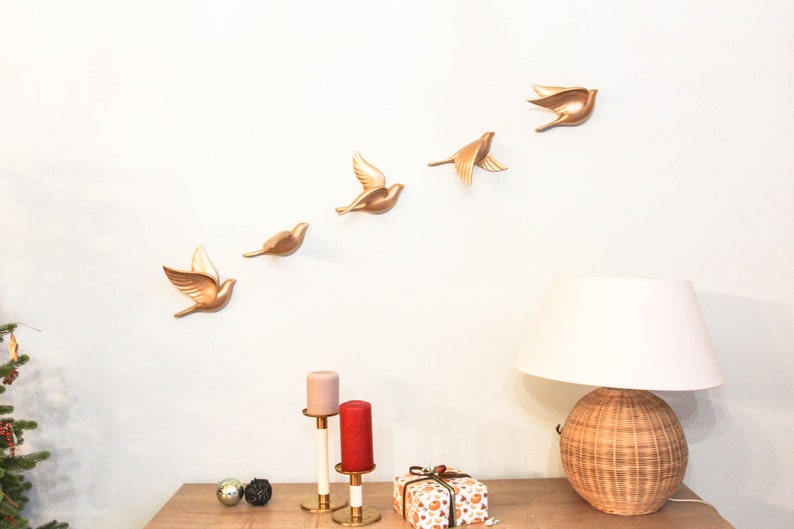 May include: A wall decoration featuring six gold-colored bird sculptures in various stages of flight. The birds are mounted on a white wall above a wooden table. A lamp with a woven base and a white shade sits on the table, along with candles and a wrapped gift.