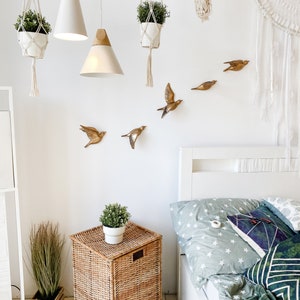 May include: A bright bedroom scene with white walls, featuring hanging plants, pendant lights, and decorative gold bird wall art. A woven basket nightstand holds a potted plant, and a bed with star-patterned bedding is visible.