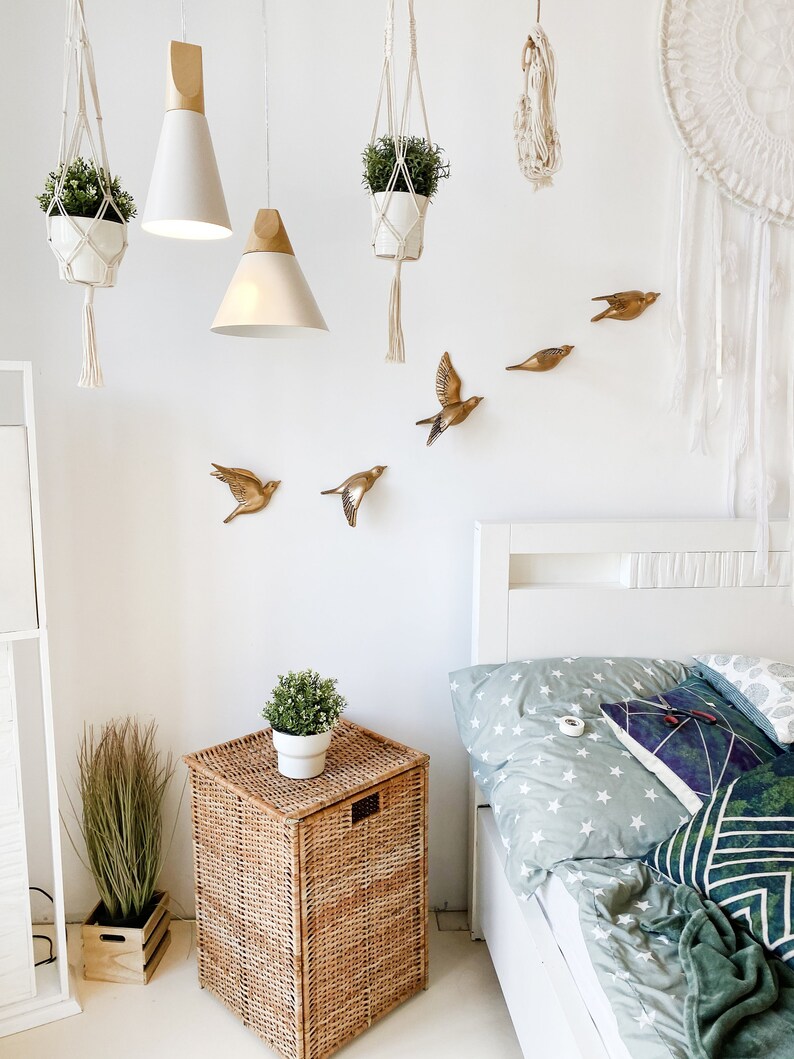 May include: A bright bedroom scene with white walls, featuring hanging plants, pendant lights, and decorative gold bird wall art. A woven basket nightstand holds a potted plant, and a bed with star-patterned bedding is visible.