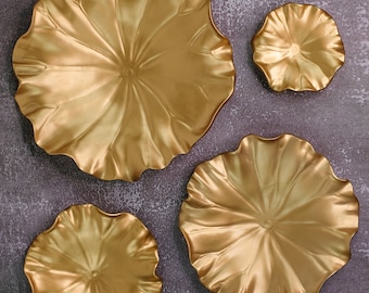 Op de afbeelding: Vier gouden decoratieve wandplaten in de vorm van lotusbloemen. De platen zijn in een verspringend patroon op een grijze achtergrond gerangschikt.