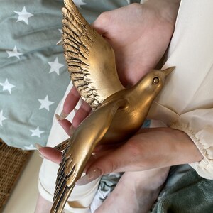May include: A golden bird sculpture with outstretched wings. The bird is held in a person's hands. The sculpture has detailed feathering on the wings and body. The bird is a decorative item, possibly for home decor.