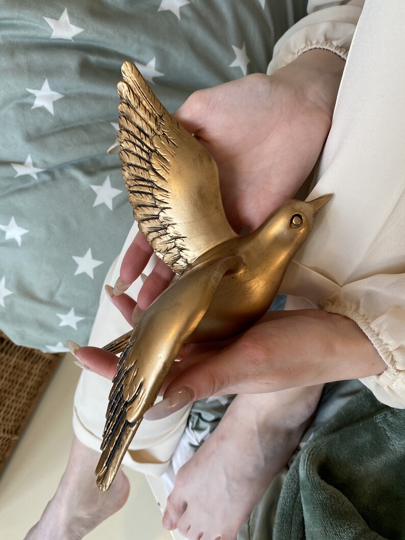 May include: A golden bird sculpture with outstretched wings. The bird is held in a person's hands. The sculpture has detailed feathering on the wings and body. The bird is a decorative item, possibly for home decor.