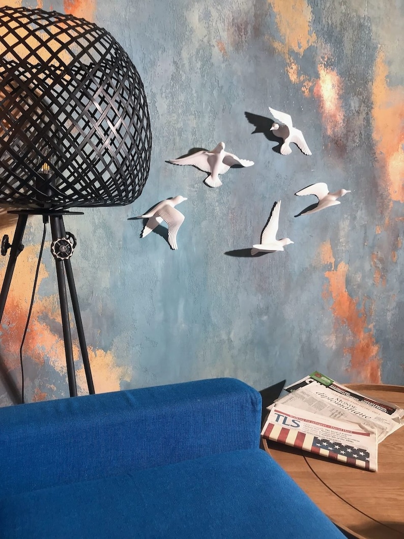 May include: A modern living room scene featuring a black geometric lamp, a blue sofa, and a wall adorned with white bird sculptures. The wall has a textured blue and orange painted design. Newspapers are on a wooden table.