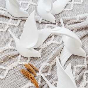May include: White ceramic bird sculptures in various poses, with wings spread or in flight. The birds are arranged on a gray, textured fabric with a white geometric pattern. Dried wheat stalks are in the foreground.