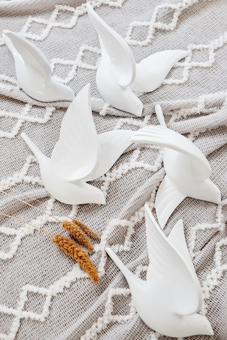 May include: White ceramic bird sculptures in various poses, with wings spread or in flight. The birds are arranged on a gray, textured fabric with a white geometric pattern. Dried wheat stalks are in the foreground.