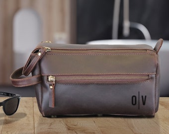 May include: A brown leather toiletry bag with a zipper closure and the initials "O/V" embossed on the front. The bag is sitting on a wooden surface next to a pair of black sunglasses, a small bottle of perfume, and a black wallet.