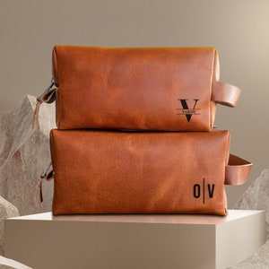 May include: Two brown leather travel bags with zipper closures and leather handles. The top bag has the initials "V|VARIN" embossed on the front. The bottom bag has the initials "O|V" embossed on the front.