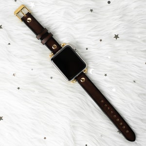 Puede incluir: Una correa de reloj de cuero marr&oacute;n con detalles dorados y tachuelas. La correa est&aacute; unida a un reloj inteligente plateado con una pantalla negra.