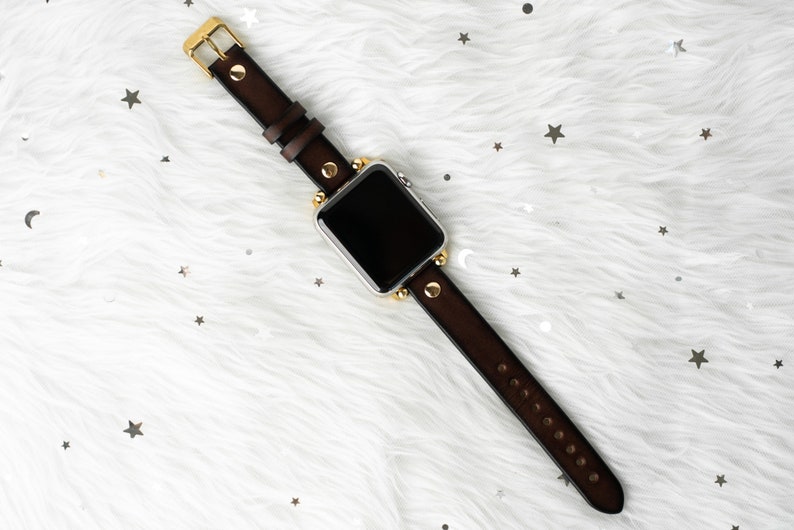 Puede incluir: Una correa de reloj de cuero marr&oacute;n con detalles dorados y tachuelas. La correa est&aacute; unida a un reloj inteligente plateado con una pantalla negra.