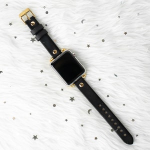 Puede incluir: Una correa de reloj de cuero negro con detalles dorados y una esfera de reloj plateada. La correa tiene un dise&ntilde;o tachonado y se muestra sobre una superficie blanca y peluda.
