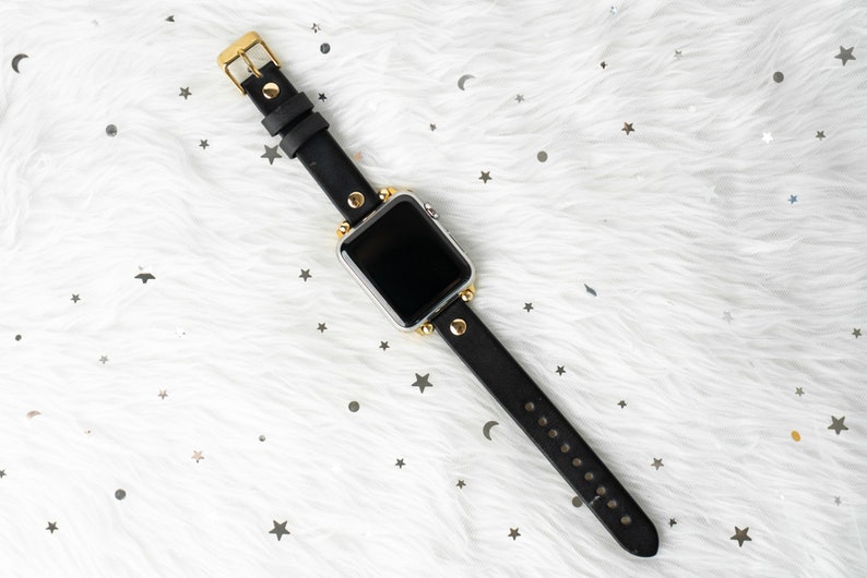 Puede incluir: Una correa de reloj de cuero negro con detalles dorados y una esfera de reloj plateada. La correa tiene un dise&ntilde;o tachonado y se muestra sobre una superficie blanca y peluda.