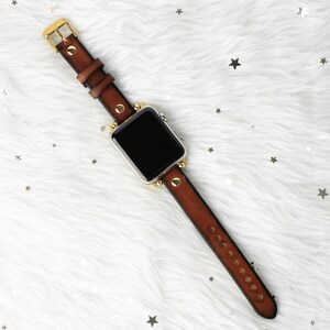 Puede incluir: Una correa de reloj de cuero marr&oacute;n con detalles dorados y una esfera negra. La correa tiene un dise&ntilde;o tachonado y se muestra sobre una superficie blanca y esponjosa.