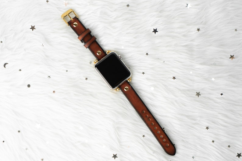Puede incluir: Una correa de reloj de cuero marr&oacute;n con detalles dorados y una esfera negra. La correa tiene un dise&ntilde;o tachonado y se muestra sobre una superficie blanca y esponjosa.