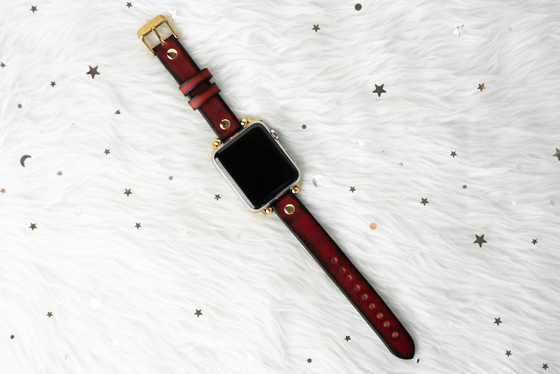 Puede incluir: Una correa de reloj de cuero rojo con detalles dorados y una esfera negra.