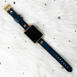 Puede incluir: Una correa de reloj de cuero azul con detalles dorados y una esfera de reloj inteligente plateada.