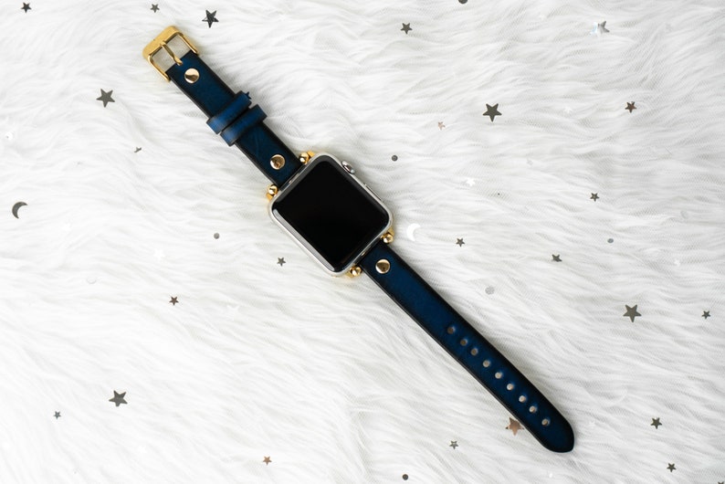 Puede incluir: Una correa de reloj de cuero azul con detalles dorados y una esfera de reloj inteligente plateada.